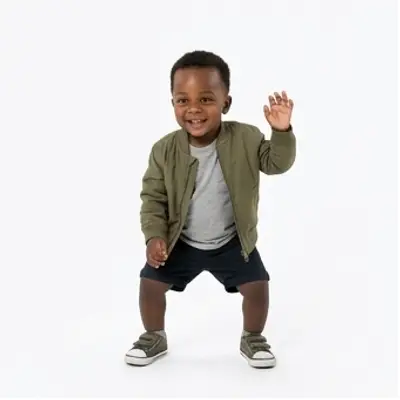 A toddler in a small olive jacket, grey t-shirt and shorts dances on a white background, arms out, then both hands in the air