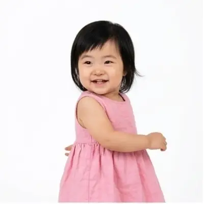 A toddler girl with a dark bob in a pink dress dances on a white background, clapping and lifting one knee