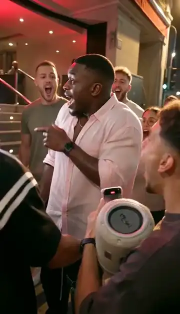 A man in a pale pink shirt raps and points at a phone held up next to a portable speaker, with a crowd filming him in front of a red-lit entrance