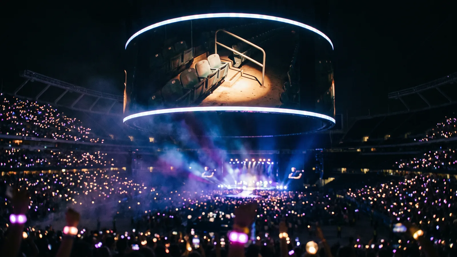 A giant round stadium jumbotron at night showing an empty railing and seats lit by a single spotlight, waiting for the next couple on the Coldplay kiss cam