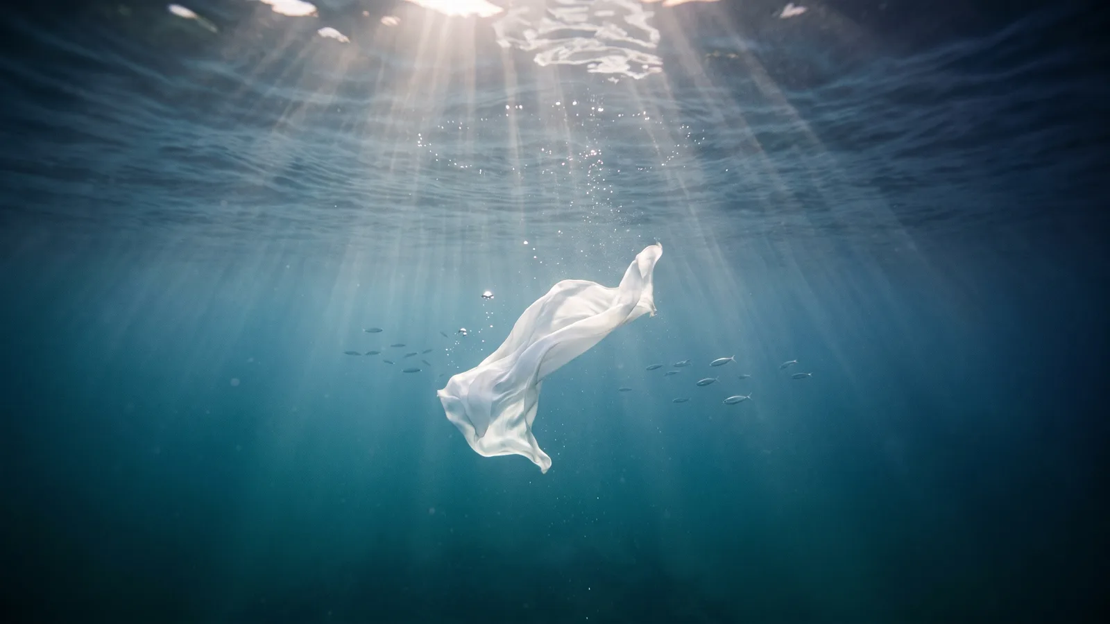 Sunbeams falling through deep blue ocean water onto a floating white silk scarf in an empty space, with bubbles rising and a school of silver fish in the background