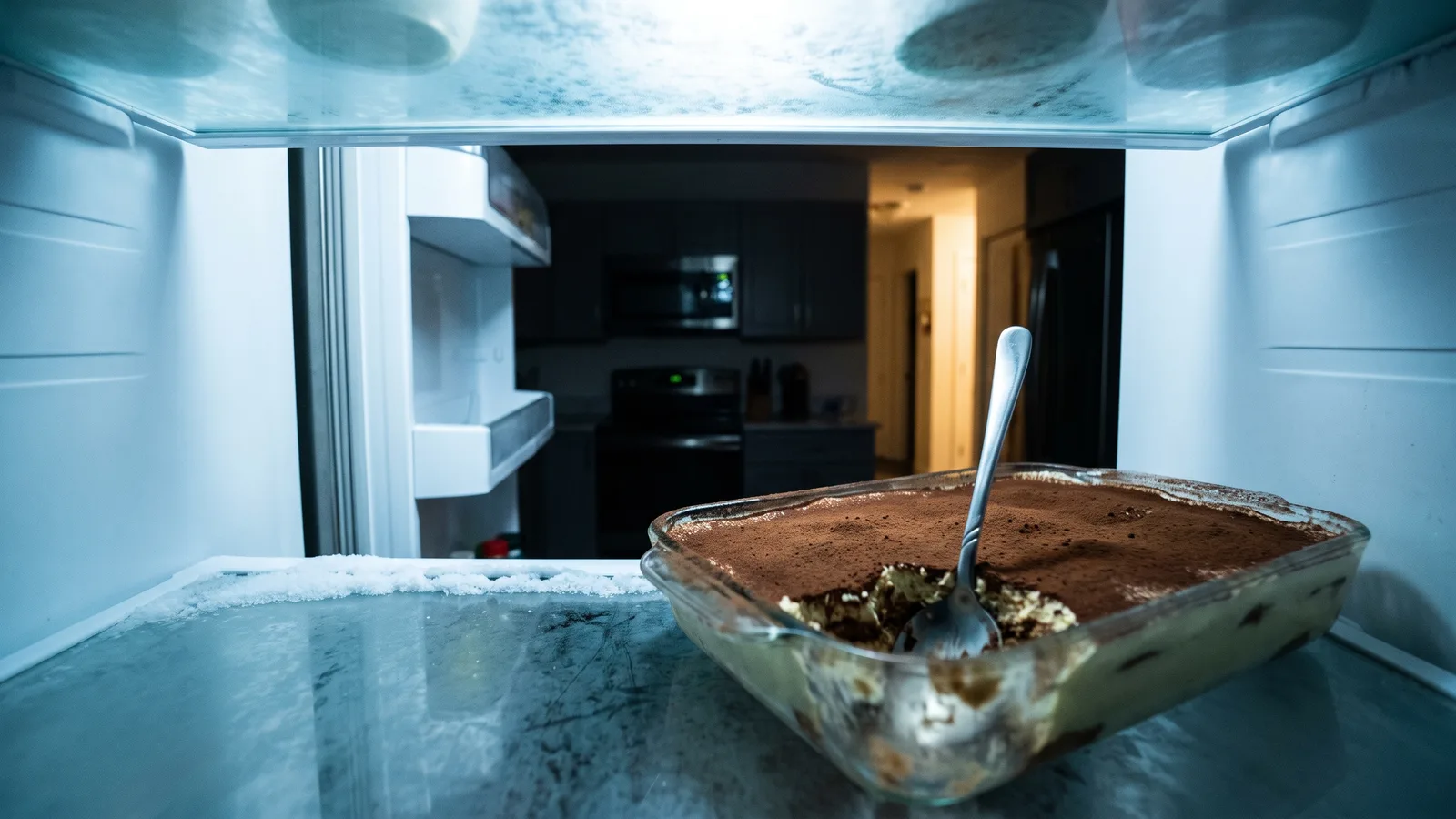A tray of tiramisu with one scoop missing and a spoon standing in it, seen from inside an open fridge looking out at a dark, empty kitchen at night