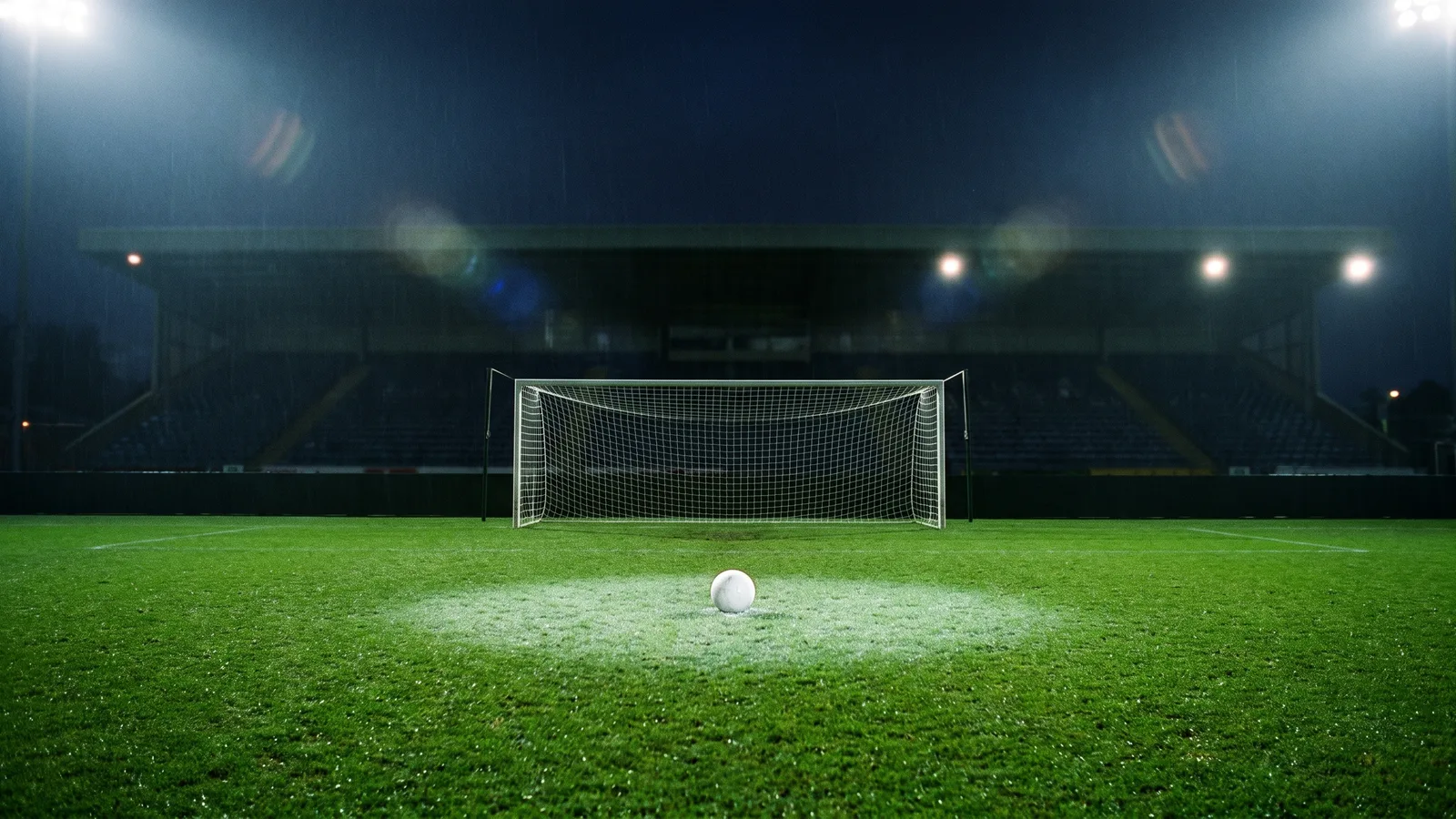 A white football on the floodlit grass in front of an empty goal in a dark, empty night stadium