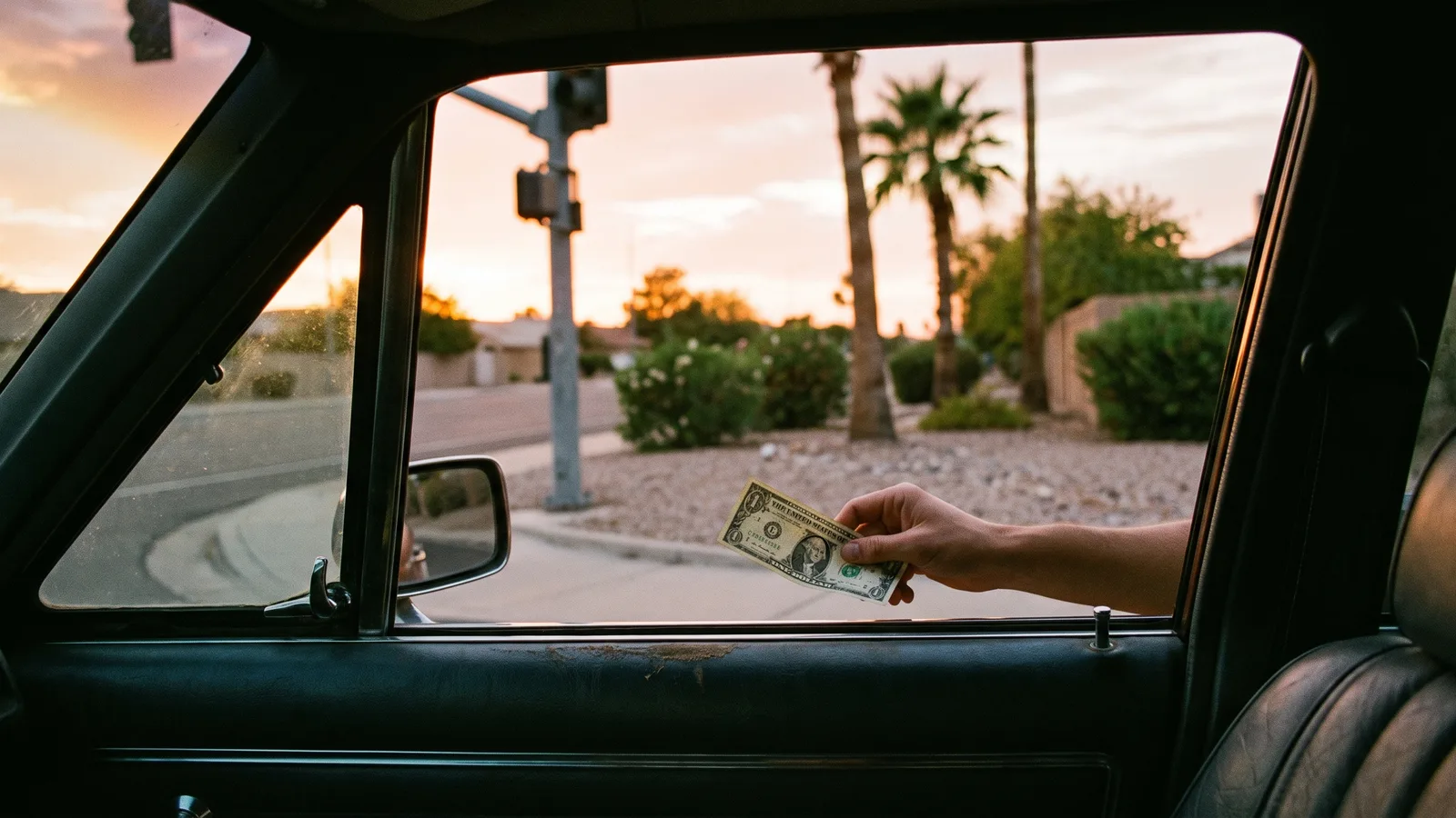 View from a car's driver's seat through the open window at an empty sunset street corner with gravel and a traffic-light pole, a hand on the window sill holding out a single one-dollar bill