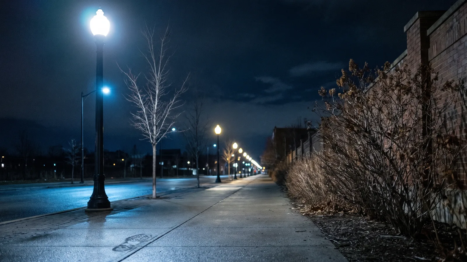 An empty night-time sidewalk under a glowing street lamp, with a scuff mark leading out of the frame on the left
