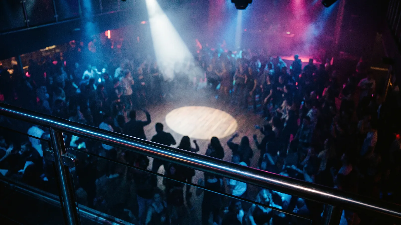 A chrome balcony railing overlooking a crowded, hazy nightclub dance floor where a single spotlight lights an empty circle in the middle of the crowd