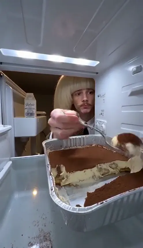 A person in pyjamas opens the fridge at night and digs a spoon straight into a tray of tiramisu, seen from inside the fridge