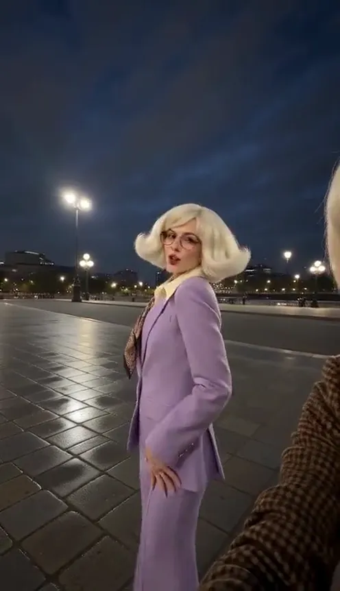 Three friends film a night-out selfie vlog on a lit city square, the one holding the phone laughing into the lens while the other two dance behind