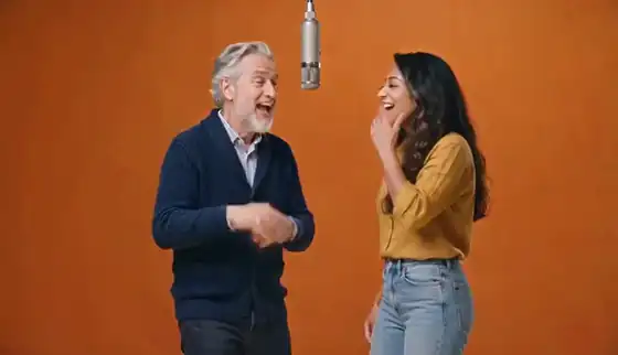 An older man in a navy cardigan and a young woman in a mustard shirt point at the camera and rap under a hanging microphone in front of an orange backdrop