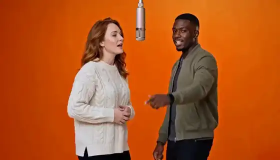 Two friends in a cream sweater and an olive jacket take turns gesturing at the camera beneath the hanging microphone