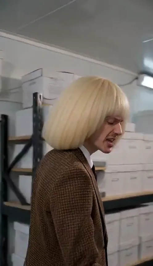 A man in a brown suit raps into the camera inside a white cold storage room lined with shelves of plain white boxes