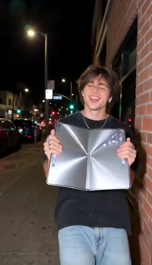 A young man on a night street in a black tee and jeans holds a phone unfolded into a giant silver tablet against his chest mid-dance, about to throw it up out of frame