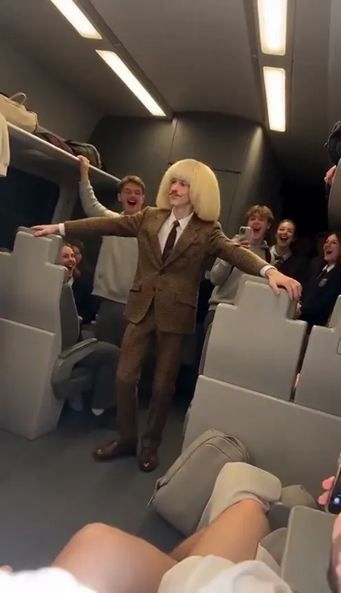 A passenger dances with arms spread wide down the aisle of a crowded train while other passengers film