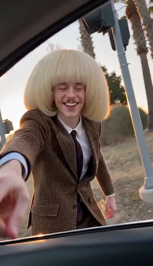 A man in a brown suit and a bob wig stands at a sunset street corner outside the open car window, grinning with a one-dollar bill in his hand