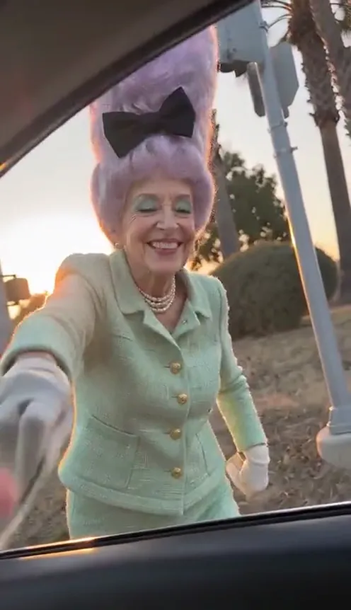 An older woman with a purple beehive and a mint-green suit does the plug walk on the gravel at sunset, dollar bill in hand, seen through the car window