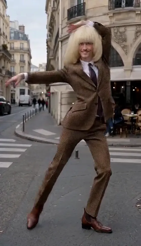 A man in a brown suit does the SaWaDiKa dance in front of a street café, palms pressed together above his head