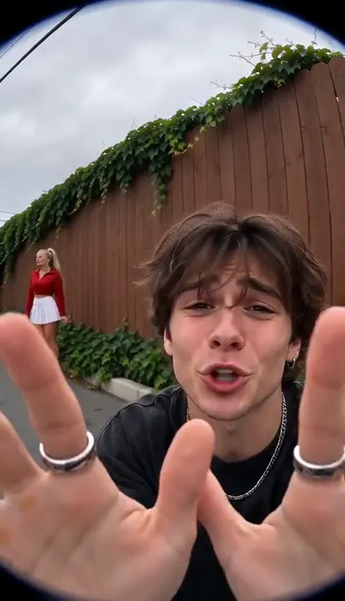 A young man in a fisheye close-up screams with both hands on his head in front of a red wooden fence with ivy while a blonde woman dances behind him
