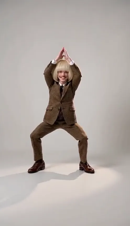 A man in a blonde bob wig and a brown suit hits the four freeze-frame poses of the I'm So In Love With You dance on a white studio backdrop