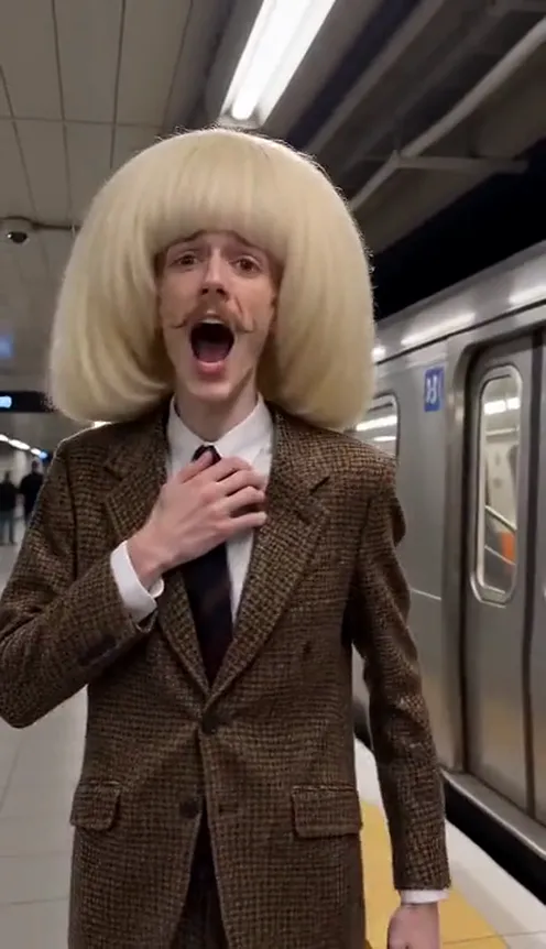 A man in a brown suit sings dramatically on a subway platform, clutching his chest