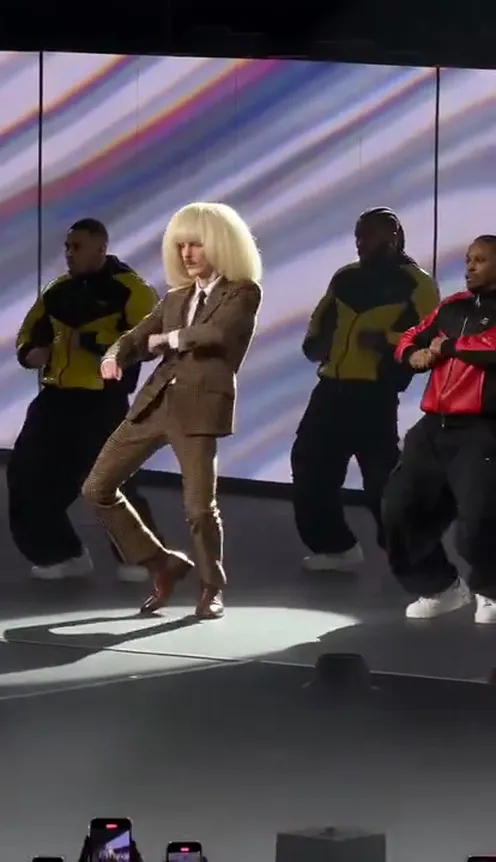 A person in their own suit leads a backup dance crew on a concert stage
