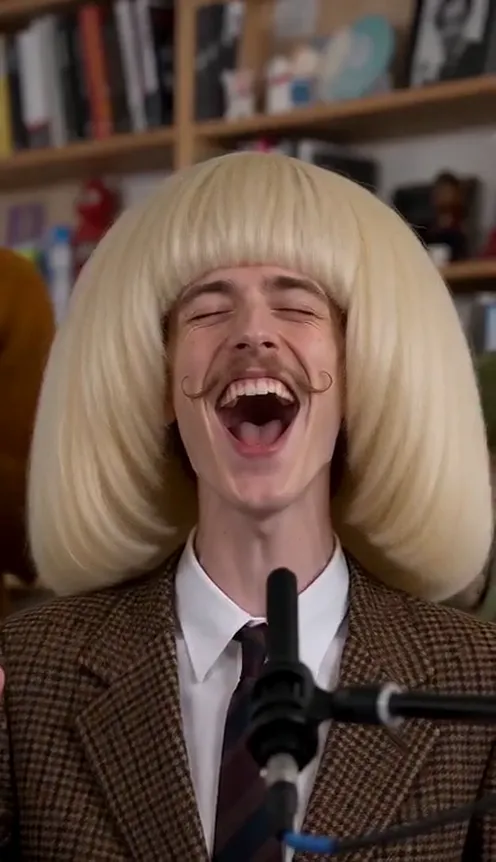 A man in a blonde wig and brown suit sings and laughs at a desk concert with a band behind him