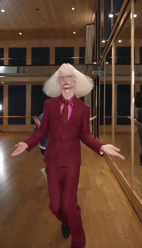 A dog standing on its hind legs dances toward the camera in a mirrored dance studio while wig-wearing dancers in suits move behind it