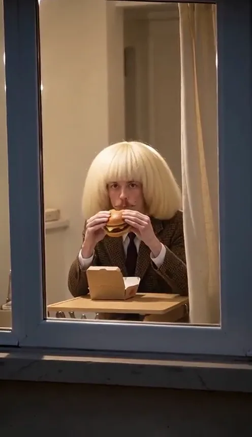 A man in a blonde bob wig and a brown vintage suit eats a burger at a table by a lit apartment window at night, staring at the camera across the street